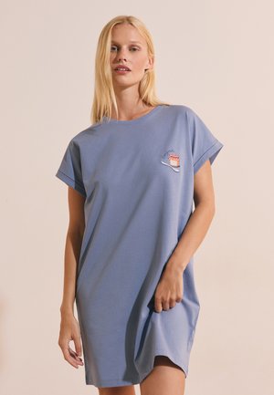 Femme blonde portant une robe t-shirt bleue ample avec un petit détail brodé de pot et de cuillère sur la poitrine.