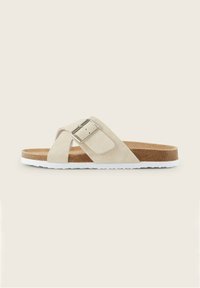 Beiga mockasin sandaler med en silverfärgad spänne, design med korsande remmar, brun fotsula och vit gummisula. Platta med en strukturerad yta.