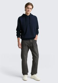 Marinblå hoodie med ribbade ärmslut, i kombination med mörkgrå jeans och vita sneakers. Minimalistisk design, avslappnad passform och enkel textur.