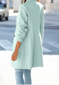 Světle zelený, přizpůsobený kabát s klopovým límcem, knoflíkovými manžetami a rovný střih, kombinovaný s modrými džínami. Měkká textura a minimalistický design.
