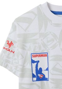 Witte shirt met een groen geometrisch patroon, met daarop de rode tekst en het logo van "Superman". Blauwe graphic die een figuur toont die een vuist omhoog steekt.