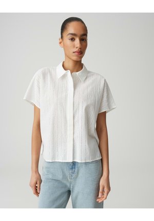 Femme portant un chemisier blanc à manches courtes avec des rayures verticales en broderie anglaise et un jean bleu clair, debout devant un fond uni.