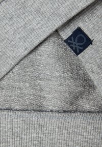 Šedá mikina s texturovaným, měkkým vnitřkem a žebrovaným lemem. Obsahuje malou tmavě modrou štítek s designem, umístěnou u výstřihu.