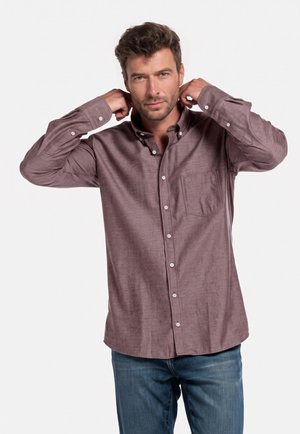 Man adjusting collar of long-sleeve brown button-up shirt, wearing blue jeans, with neutral expression against plain white background.