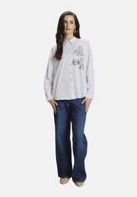 Camisa blanca de botones con bordado floral en el lado izquierdo, combinada con jeans de pierna ancha azules y zapatos beige. Textura de tela suave.