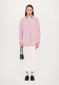 Chemise oversize rayée rose et blanche, à boutons, avec des manches longues. Portée sur une jupe blanche, assortie à des mocassins noirs et un petit sac à main.