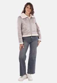 Giacca grigia in finta shearling con colletto e polsini bianchi, chiusura frontale con zip, due tasche laterali, abbinata a jeans a vita alta e sneakers bianche.