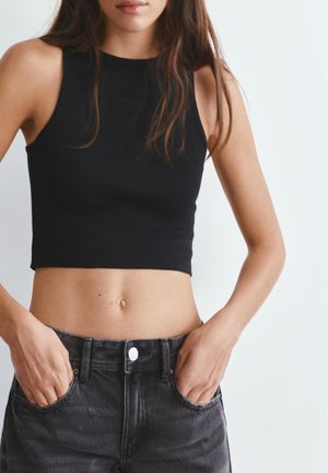 Kvinde iført sort, ærmeløs cropped top og højtaljede, falmede sorte jeans med hænderne i lommerne mod ensfarvet baggrund.