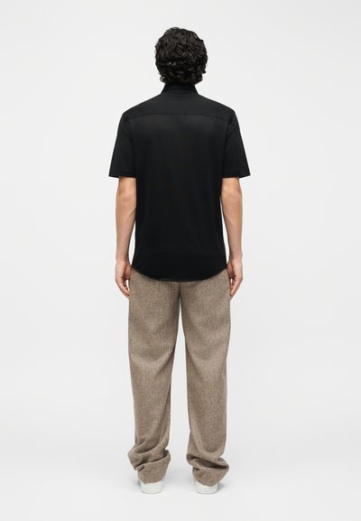 Άνδρας με σγουρά μαλλιά που φοράει μαύρο κοντομάνικο πουκάμισο και φαρδιά μπεζ παντελόνια, στέκεται γυρισμένος με την πλάτη σε μονόχρωμο φόντο.