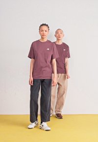Deux mannequins se tiennent sur un sol jaune. Le mannequin de devant porte un t-shirt bordeaux avec un logo, un pantalon sombre et des baskets blanches. Le deuxième mannequin porte un t-shirt bordeaux assorti et un pantalon beige.