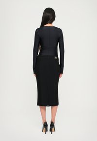 Top preto de mangas compridas, design ajustado, combinado com uma saia lápis preta com uma racha nas costas e costura contrastante. Usado com sapatos de salto pretos.