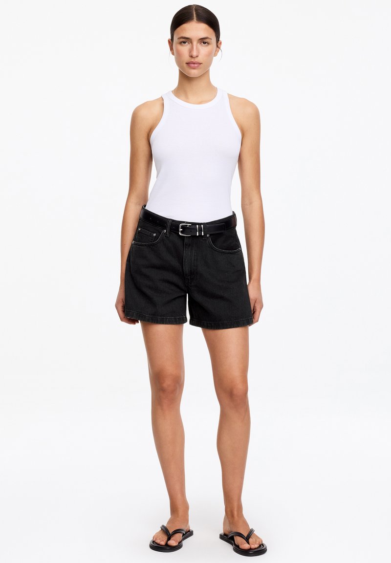 Femme portant un débardeur blanc, un short en jean noir ceinturé et des tongs noires, debout sur fond blanc.