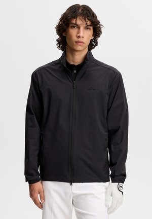 Man met een zwart jack met ritssluiting, witte broek en een witte golfhandschoen aan zijn rechterhand, staand tegen een effen achtergrond.