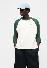 Άνδρας που φοράει λευκό και πράσινο μπλουζάκι μπέιζμπολ της Nike, μαύρο παντελόνι και ανοιχτό μπλε καπέλο, στέκεται με τα χέρια στις τσέπες κοιτώντας πλάγια.