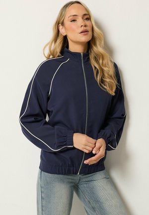 Frau mit langen blonden Haaren, die eine marineblaue Jacke mit Reißverschluss und weißen Paspeln sowie hellblaue Jeans trägt, steht vor einem hellen Hintergrund.