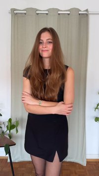 Čierne mini šaty s bočným rozparkom a krátkymi rukávmi, doplnené priehľadnými čiernymi pančuchami. V pozadí svetlo zelený záves a izbové rastliny.