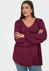 Bordeauxrote Bluse mit voluminösen Ärmeln, V-Ausschnitt und strukturierten Schulterdetails. Glatter Stoff mit lockerem Schnitt.