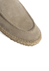 Beige suède instapper met een ronde neus, voorzien van een gevlochten jute zool en contrasterende stiksels langs de rand. Over het geheel een gladde textuur.