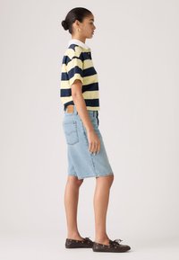 Γυναίκα που στέκεται πλαγίως φορώντας μπλε ναυτικό και ανοιχτό κίτρινο ριγέ πόλο, φούστα από ανοιχτό τζιν μήκους έως το γόνατο και σκούρα καφέ μοκασίνια.