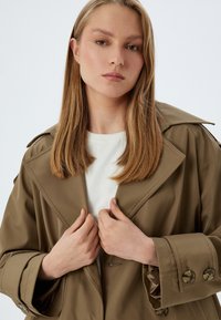 Abrigo trench color caqui con solapas anchas y puños abotonados, combinado con una camisa blanca. Tejido suave con un ligero brillo y detalles de costura intrincados.