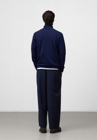 Navy Pullover-Sweater mit hohem Kragen und gerippten Bündchen, kombiniert mit locker sitzenden navyblauen Hosen und dunklen Schuhen, von hinten betrachtet.