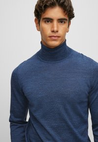 Tumši zils augsto apkakliņu džemperis, izgatavots no teksturētas adīšanas, ar augstu apkakli un garām piedurknēm.