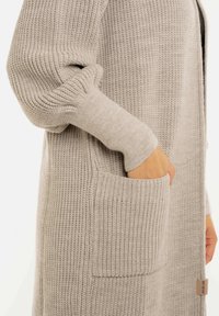 Hand gestoken in de voorzak van een beige chunky gebreide cardigan met geribbelde boorden en gestructureerde steken.