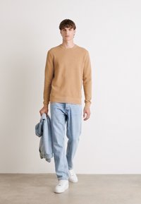 Maglione beige a trama sopra jeans in denim azzurro chiaro, abbinato a sneaker bianche e mentre tiene una giacca di denim azzurro chiaro.