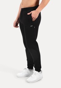 Sorte joggingbukser med tilspidset pasform, elastisk taljebånd, to sidelommer og små mærkedetaljer, kombineret med hvide sneakers.