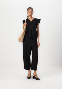 Top corto nero a maniche corte con dettagli arricciati abbinato a pantaloni neri larghi. La modella tiene in mano una borsa di paglia intrecciata e indossa décolleté nere con cinturino.
