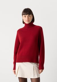 Rød turtleneck genser laget av mykt, ribbet materiale, med en avslappet passform og lange ermer, sammen med et kremfarget plisséskjørt.