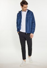 Marineblaues kapuzenjacke mit einem Reißverschluss vorne, zwei Brusttaschen und elastischen Bündchen; kombiniert mit schwarzen Slim-Fit-Hosen und weißen Turnschuhen.