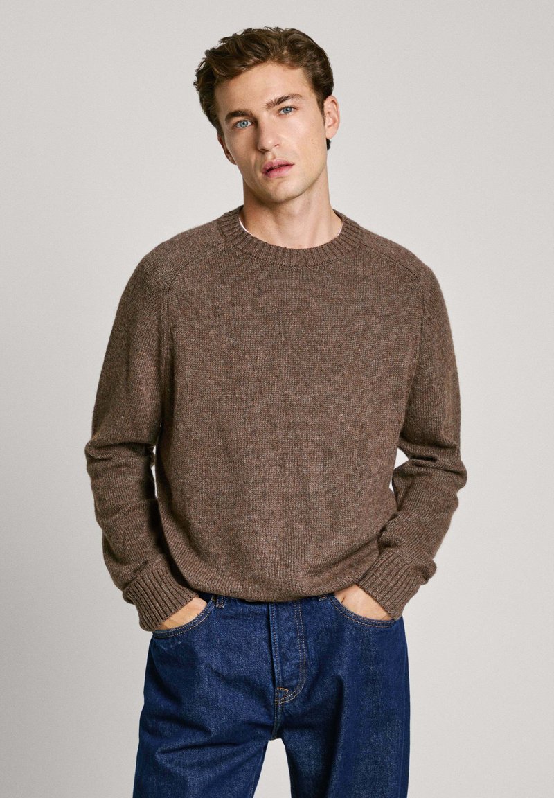 Suéter de punto marrón con cuello redondo, puños acanalados y dobladillo. Combinado con unos vaqueros de denim azul, con un corte relajado y bolsillos visibles.