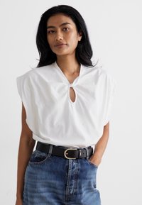 White cotton top with wide sleeves and a keyhole neckline, paired with high-waisted blue denim jeans and a black belt with a gold buckle.