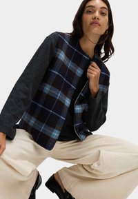 Kratkowana kamizelka w ciemnoniebieskim i brązowym wełnianym materiale z szarymi rękawami, zestawiona z szerokimi spodniami w kolorze kremowym i czarnymi butami, o swobodnym kroju.