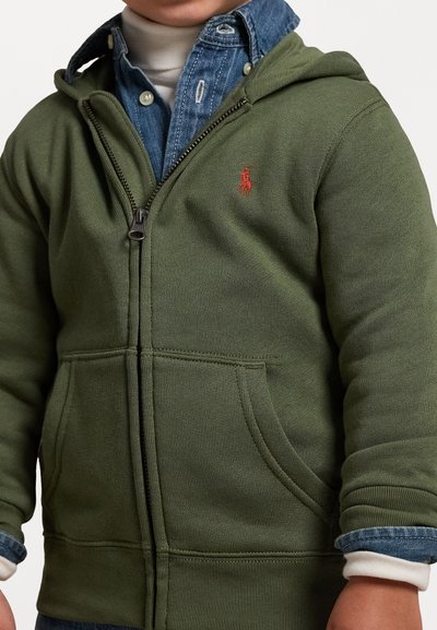 Zelená mikina na zip s přední kapsou, žebrovanými manžetami a malým červeným logem. Nositá přes modrou džínsovou košili. Měkká, texturovaná tkanina.