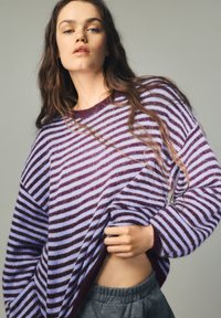 Pruhovaný sveter v purpurovej a svetlomodrej farbe, vyrobený z mäkkého materiálu. Boxy strih so širokými rukávmi, skombinovaný so sivými voľnými nohavicami.