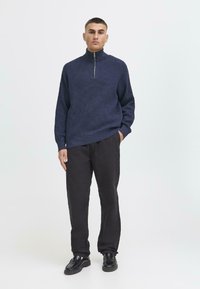 Marineblauer Reißverschluss-Pullover aus strukturiertem Stoff, mit weitem Kragen und langen Ärmeln, kombiniert mit schwarzen Hosen und schwarzen Schuhen. Minimalistisches Design.