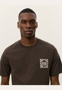 Camiseta de algodón marrón de manga corta y cuello redondo. Cuenta con un estampado de bolsillo gráfico cuadrado en un patrón de cuadros en blanco y negro.