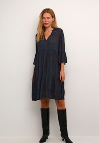 Abito blu navy con scollo a V, design fluido e maniche a campana; presenta un motivo sottile e una gonna a strati. Indossato con stivali neri al ginocchio.