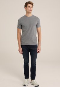 Camiseta gris de manga corta con cuello redondo, combinada con vaqueros azul oscuro y zapatillas blancas, presentada sobre un fondo neutral.