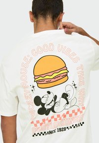 Camiseta de algodón blanca con un gráfico de una hamburguesa encima de "MICKEY MOUSE: BUENAS VIBRACIONES POR AQUÍ" y "OBYDO HAMBRIENTO DESDE 1928" en colores brillantes.