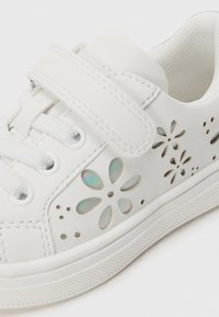 Vita sneakers med perforerade blommönster, syntetiskt material, snörning och kardborreknäppning, texturerad gummisula och subtila iriserande detaljer.