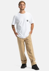 Witte katoenen t-shirt met een borstzak, voorzien van een zwart logo, gecombineerd met lichtbeige broek en zwarte sneakers. Casual pasvorm.
