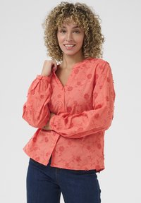 Blusa di corallo con ricami floreali, chiusura frontale con bottoni, scollatura a V e maniche a sbuffo. Tessuto morbido con dettagli testurizzati e vestibilità rilassata.