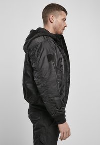 Giacca bomber nera con cappuccio, polsini a costine, zip frontale e patch in velcro sulla manica. Texture opaca, design leggero e silhouette casual.