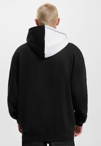 Zwarte hoodie met een witte capuchon, gemaakt van zacht materiaal, ontspannen pasvorm, ribbelmanchetten en -zoom, en een contrasterende stikseldetail langs de rand van de capuchon.