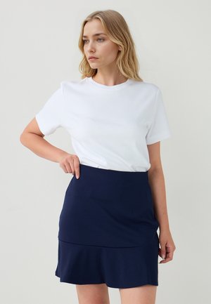 Femme aux cheveux blonds portant une chemise blanche à manches courtes et une jupe bleu marine, pointant vers la taille, fond neutre.