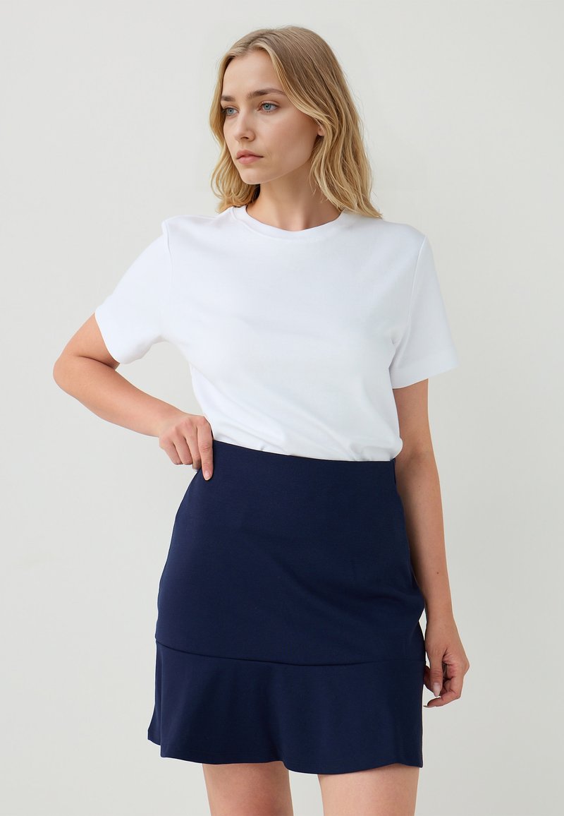 Vrouw met blond haar, gekleed in een witte shirt met korte mouwen en een marineblauwe rok, wijst naar de taille, neutrale achtergrond.