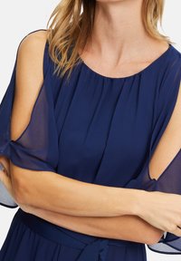 Femme portant une robe bleu marine avec des manches épaules transparentes, bras croisés à la taille et cheveux blonds clairs partiellement visibles.